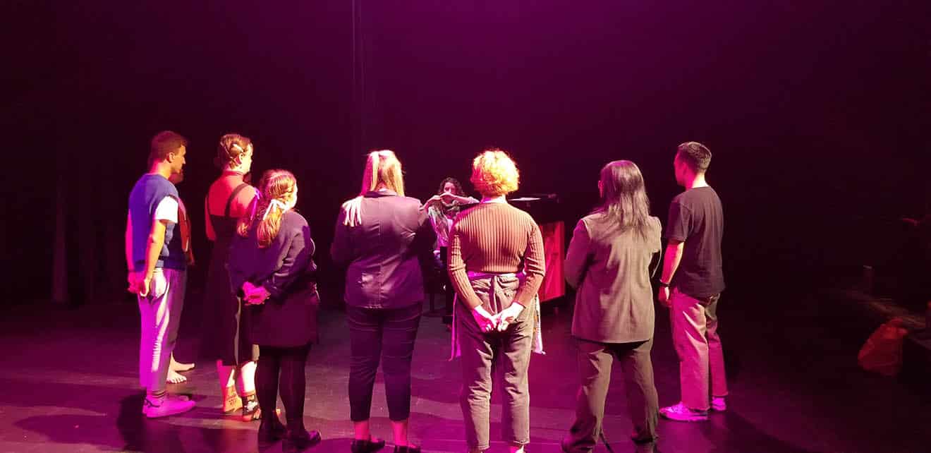 Students standing in a semi-circle on a stage looking towards a person at a piano talking to them.